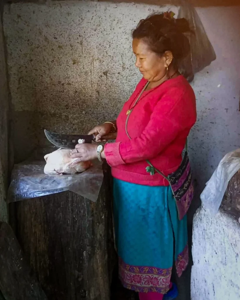A woman in a red sweater and blue skirt stands in a rustic room, using a large knife to cut a piece of raw meat on a wooden stump covered in plastic.