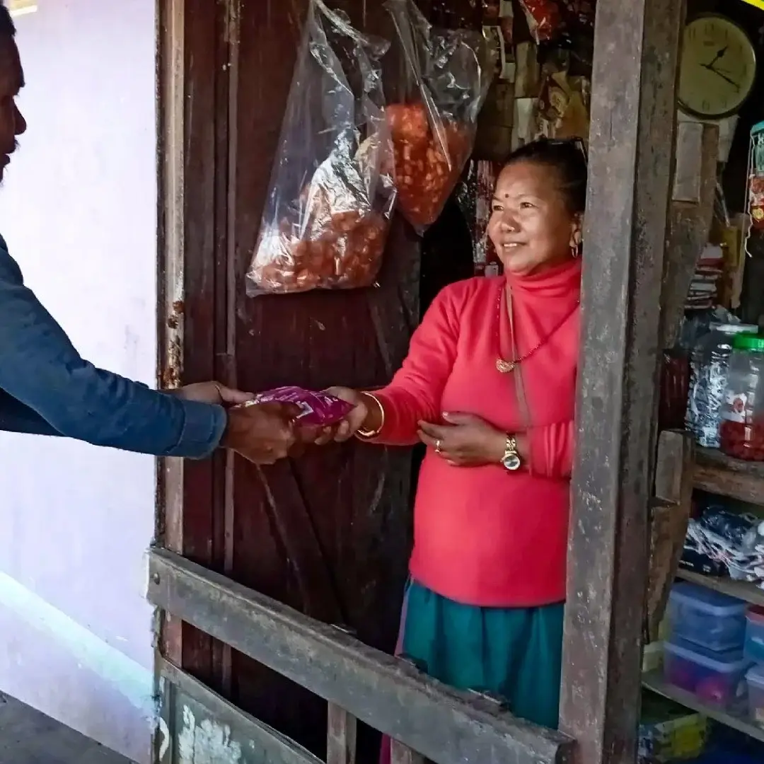 A man in a blue sweater hands a small purple snack bag to a smiling woman in a red turtleneck standing inside a small, well-stocked village shop in Nepal.