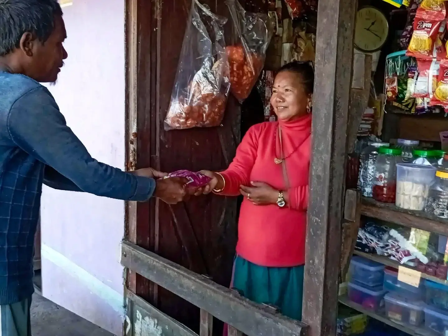 A man in a blue sweater hands a small purple snack bag to a smiling woman in a red turtleneck standing inside a small, well-stocked village shop in Nepal.