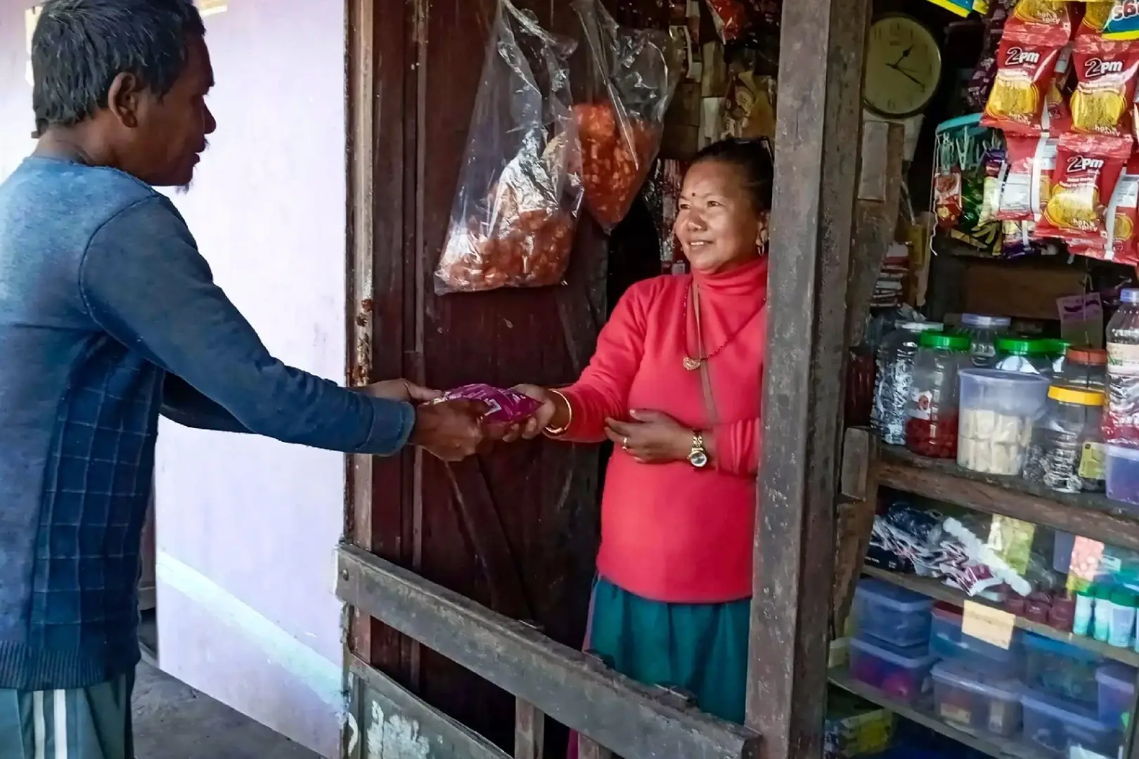 A man in a blue sweater hands a small purple snack bag to a smiling woman in a red turtleneck standing inside a small, well-stocked village shop in Nepal.