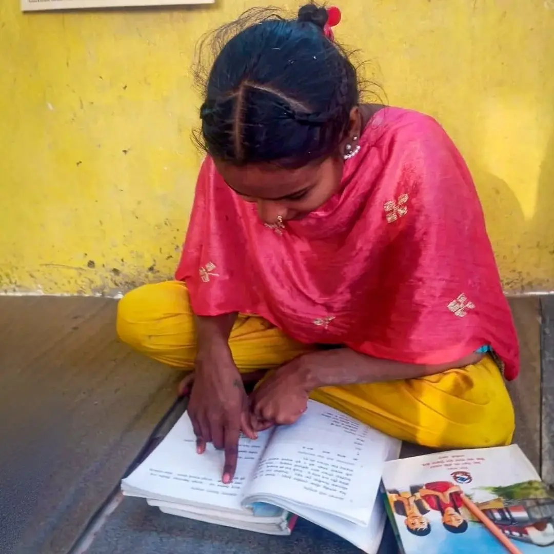 Young girl in a pink shawl and yellow pants studies an open workbook on a mat outdoors, focused beside colorful books against a yellow wall.