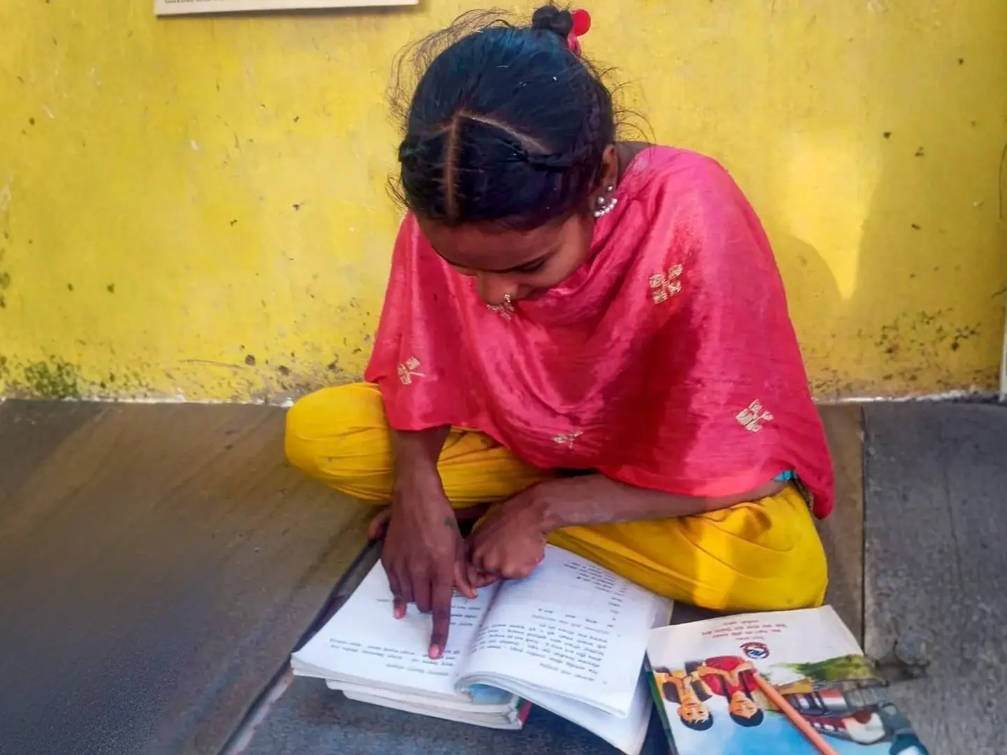 Young girl in a pink shawl and yellow pants studies an open workbook on a mat outdoors, focused beside colorful books against a yellow wall.