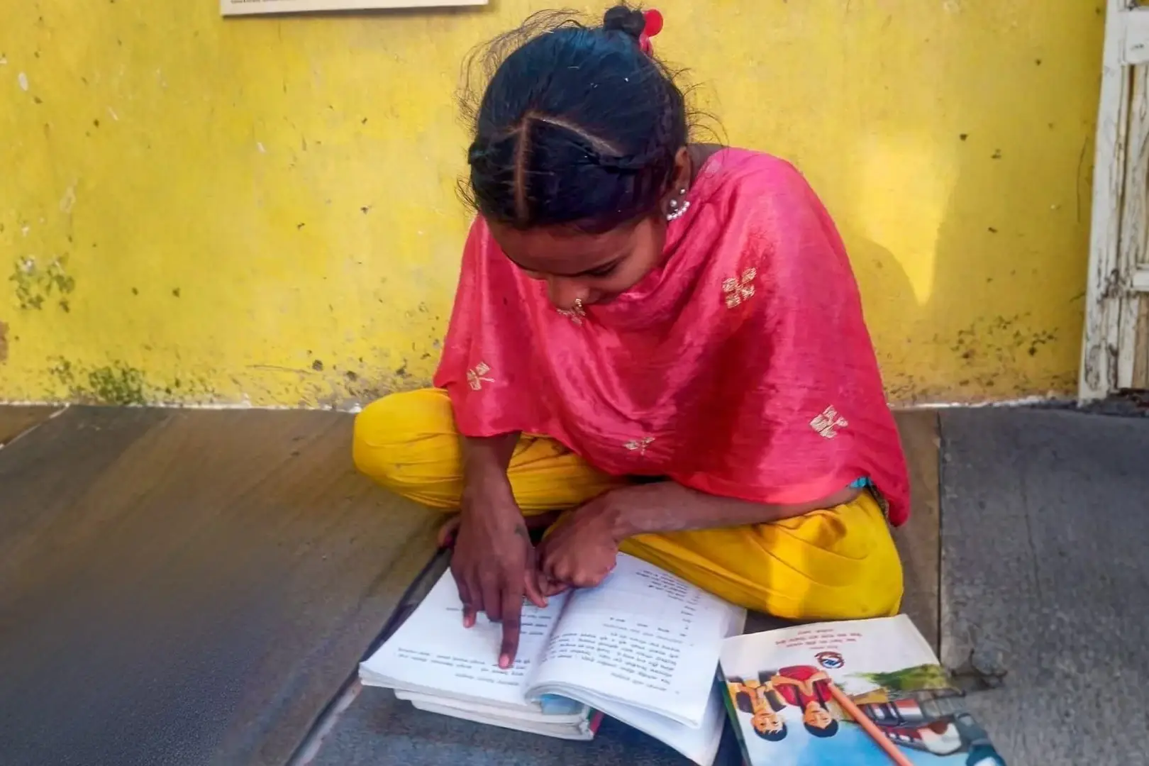 Young girl in a pink shawl and yellow pants studies an open workbook on a mat outdoors, focused beside colorful books against a yellow wall.
