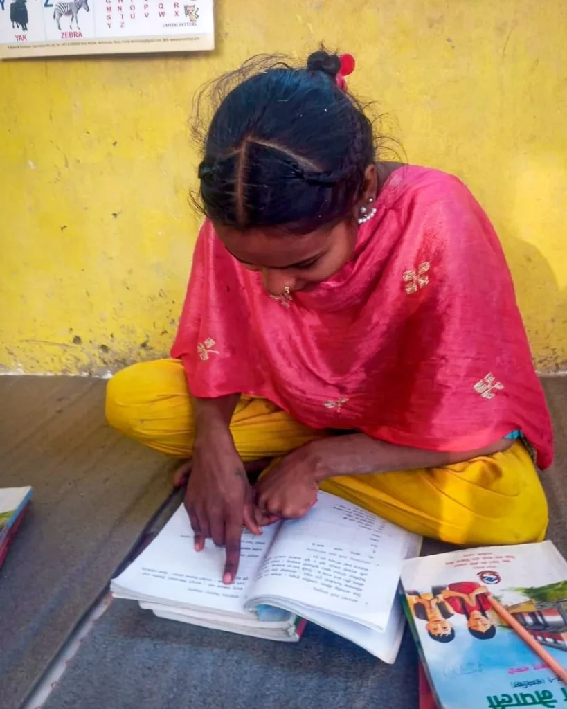 Young girl in a pink shawl and yellow pants studies an open workbook on a mat outdoors, focused beside colorful books against a yellow wall.