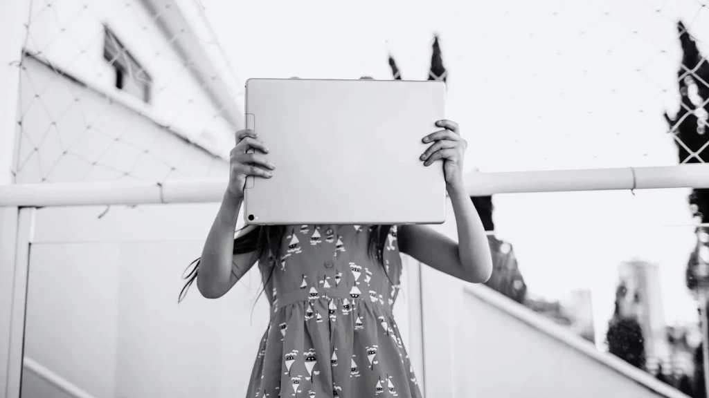 Black-and-white image of a young girl holding a tablet over her face outdoors, creating a mysterious, quiet, mood.