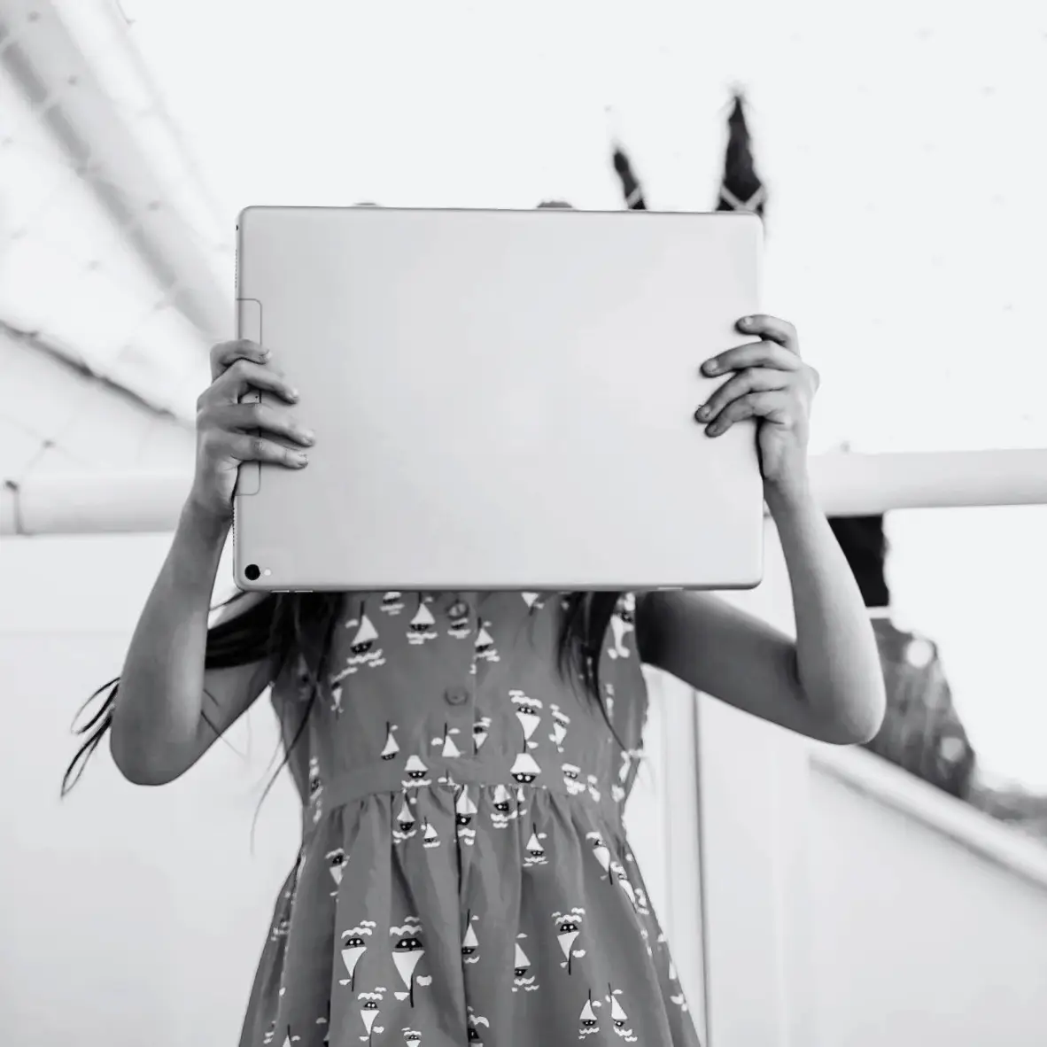 Black-and-white image of a young girl holding a tablet over her face outdoors, creating a mysterious, quiet, mood.