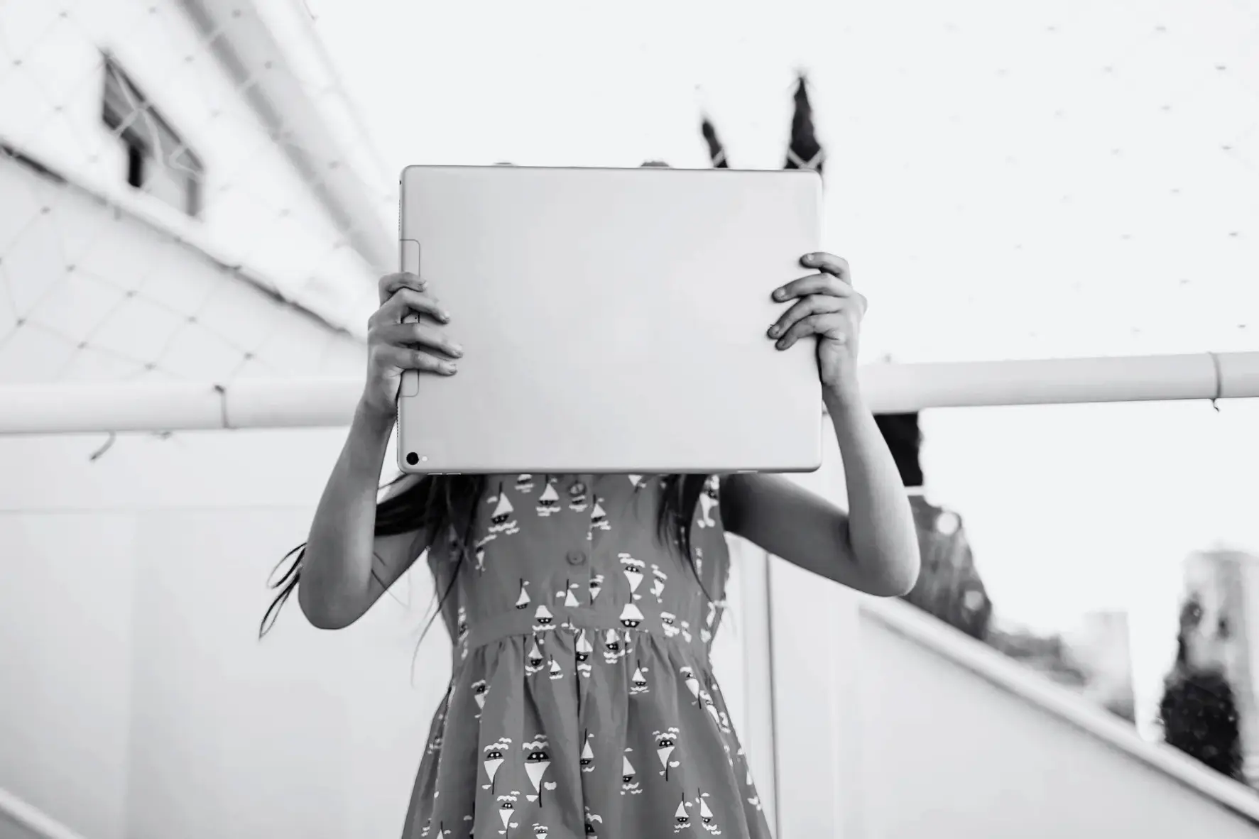 Black-and-white image of a young girl holding a tablet over her face outdoors, creating a mysterious, quiet, mood.