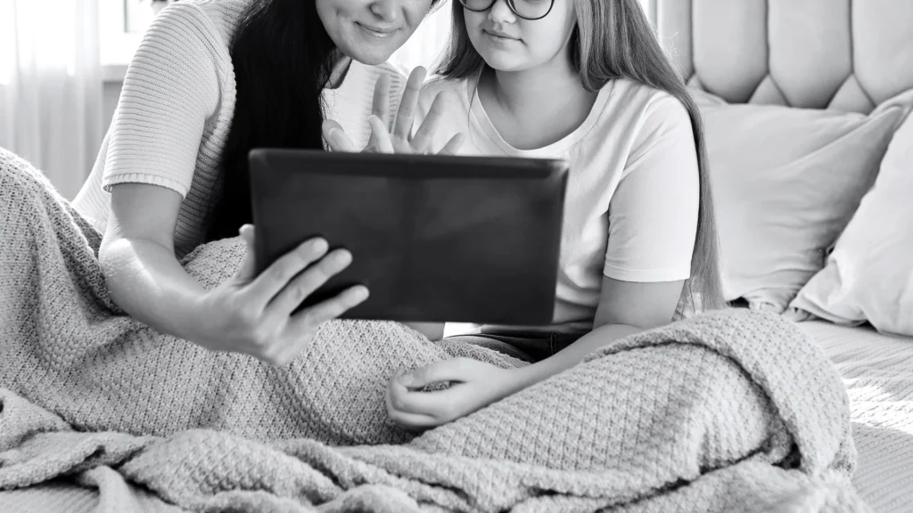 A mother and daughter sit cozily under a knitted blanket on a bed, smiling as they look together at a tablet, creating a warm family moment.