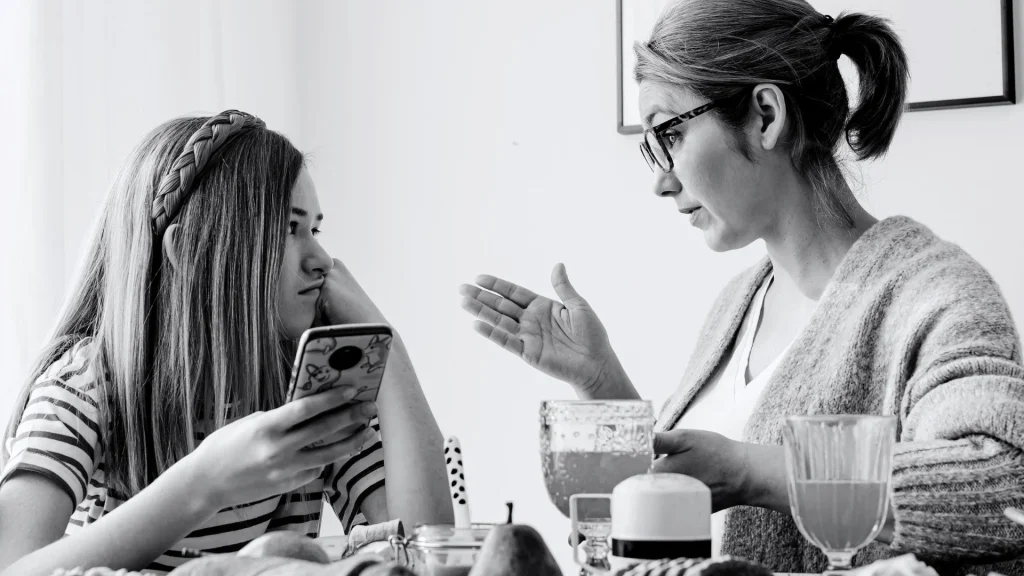 A woman and her daughter sit at a table, engaged in conversation while the daughter is holding a phone.
