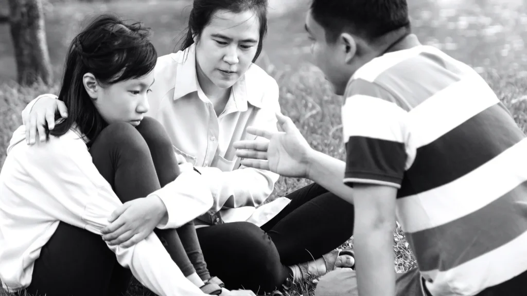A man and woman sit on the grass, comforting their daughter between them.