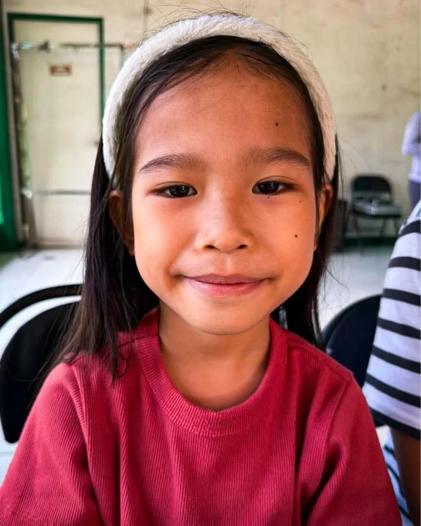 A Filipino girl with long black hair in a red top and white headband sits indoors.