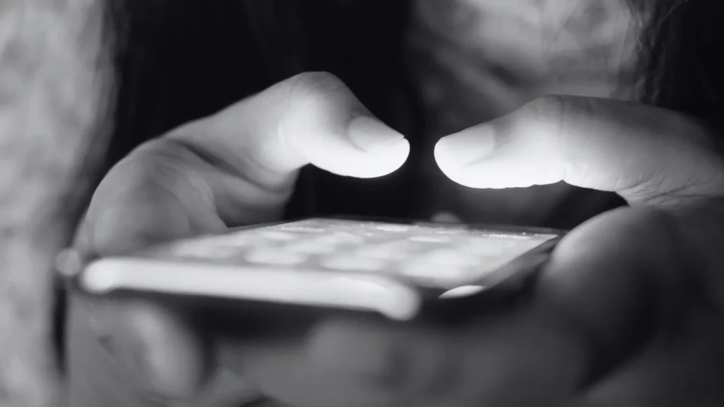 A close-up black and white shot of two thumbs typing on a glowing smartphone screen, with the light illuminating the tips of the fingers in the dark.