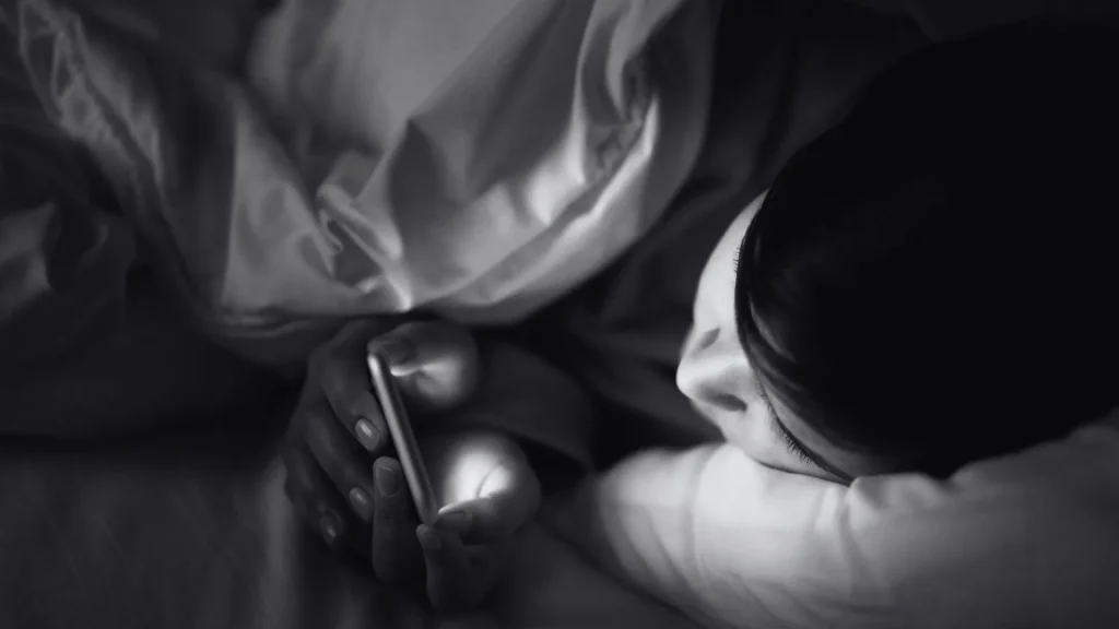 A person lying in bed under a white blanket in the dark, holding a glowing smartphone that illuminates their face and hands in black and white.