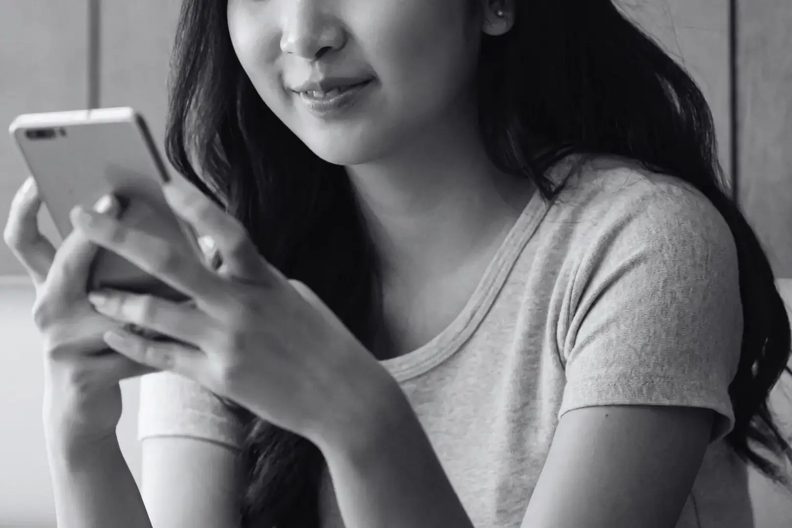 A black and white close-up of a smiling young woman with long dark hair holding a smartphone in both hands while looking down at the device screen.