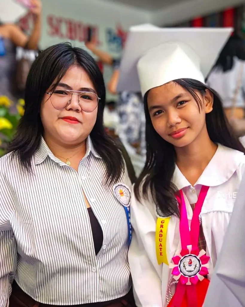 Two smiling women in the Philippines pose at a graduation ceremony; a graduate in a white cap and pink sash stands beside a woman in glasses and a striped shirt.
