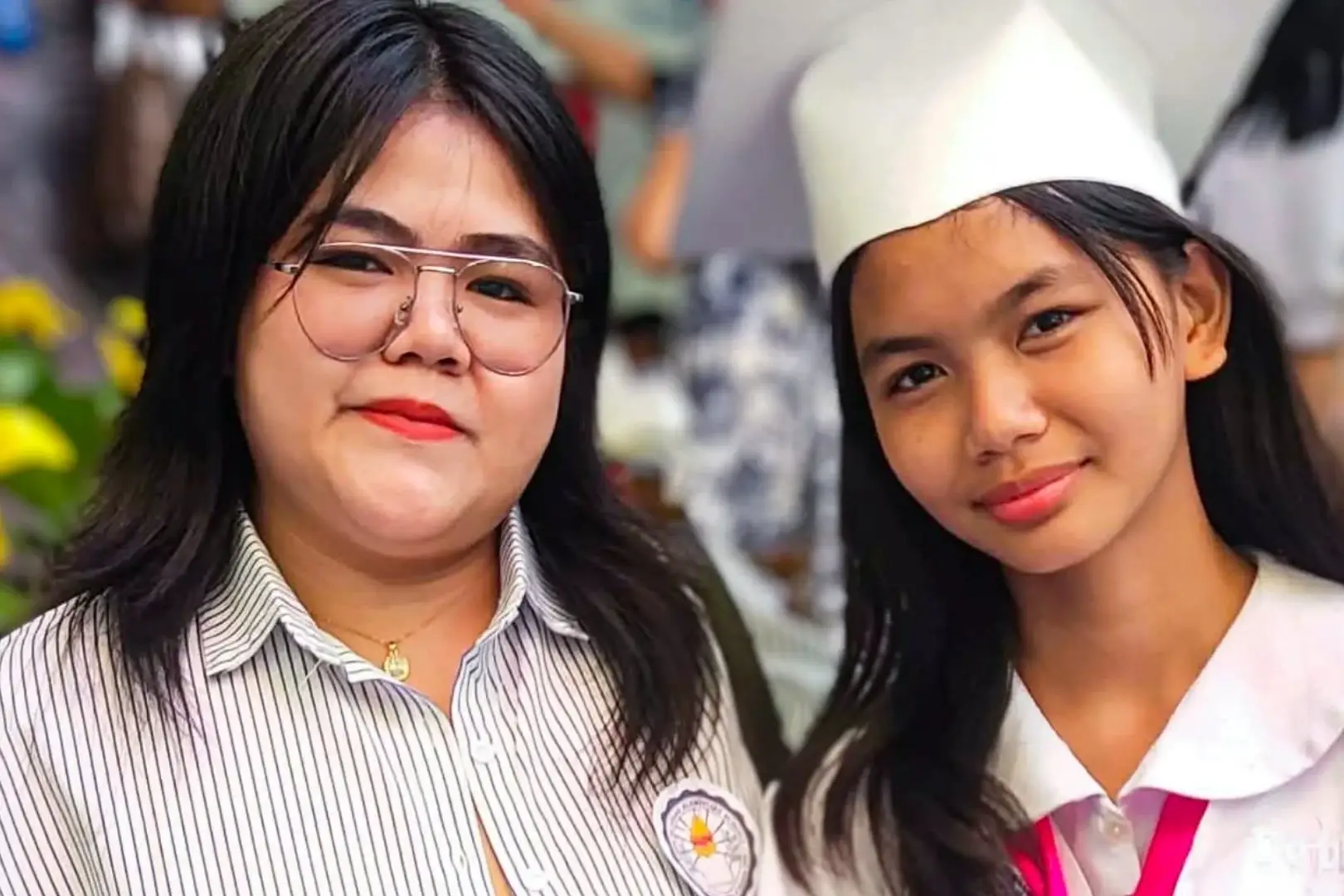 Two smiling women in the Philippines pose at a graduation ceremony; a graduate in a white cap and pink sash stands beside a woman in glasses and a striped shirt.