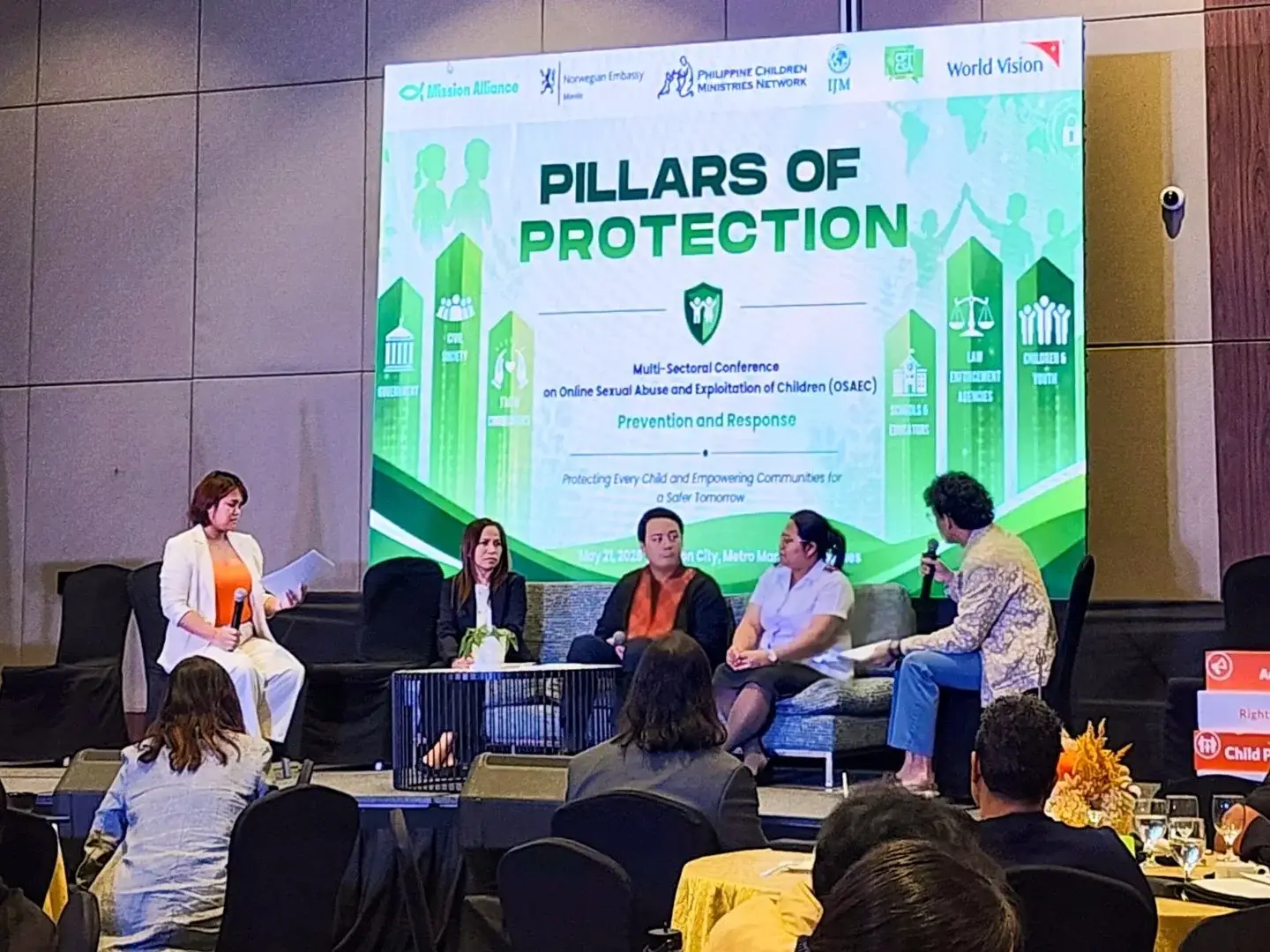 A panel of five speakers sits on a stage in front of a large screen titled Pillars of Protection at a conference about child safety and protection.