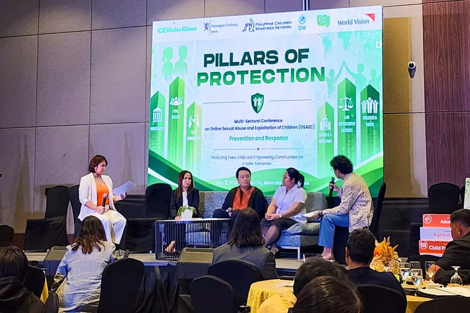 A panel of five speakers sits on a stage in front of a large screen titled Pillars of Protection at a conference about child safety and protection.