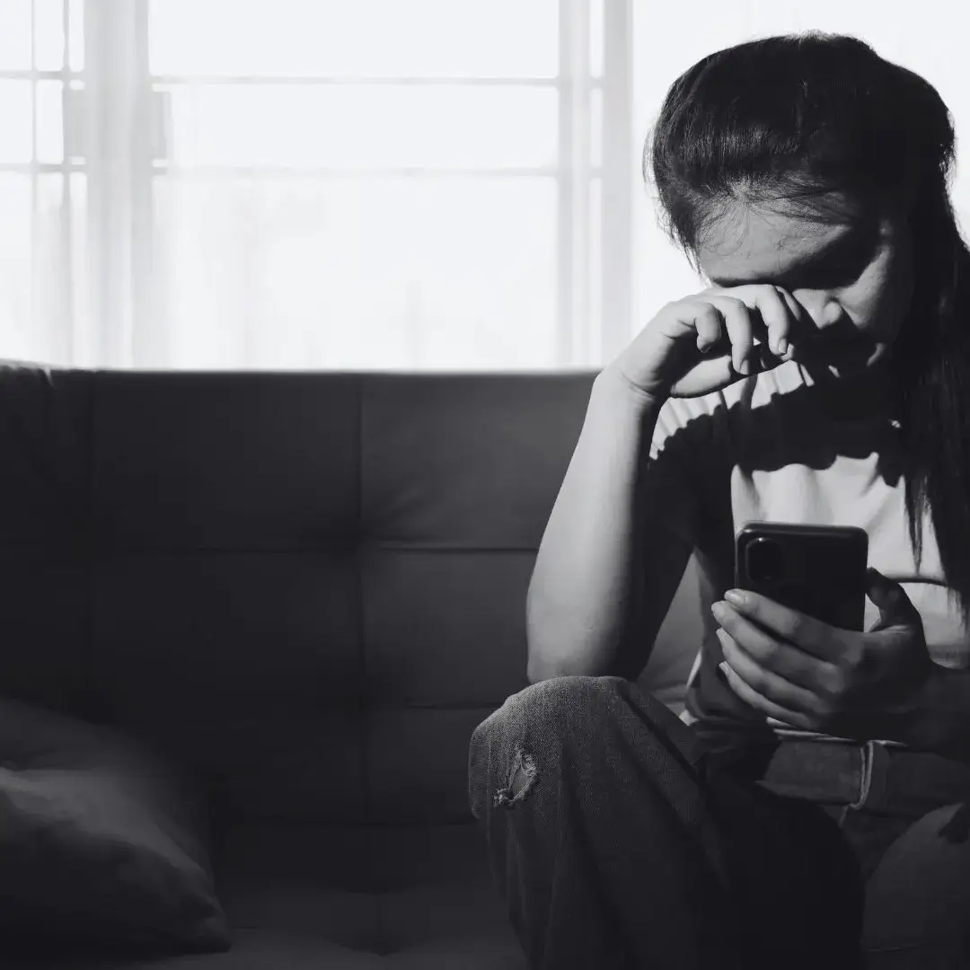 Black-and-white photo of a young woman sits alone on a dark sofa, wiping her eyes while holding a phone; bright window light contrasts with her sadness and isolation.