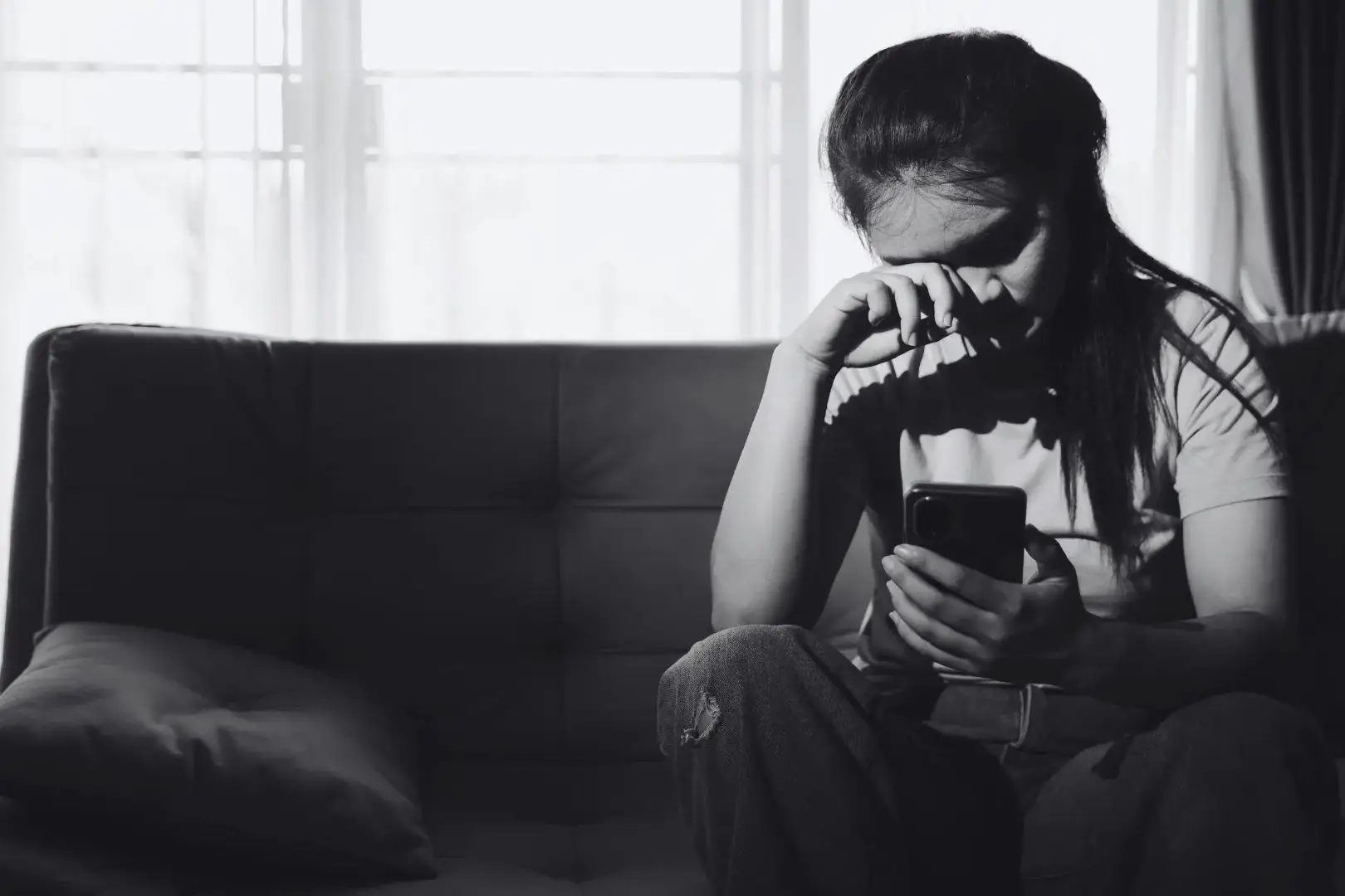 Black-and-white photo of a young woman sits alone on a dark sofa, wiping her eyes while holding a phone; bright window light contrasts with her sadness and isolation.
