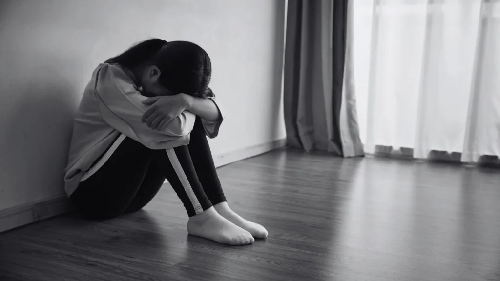 Black-and-white photo of a person sitting alone on a wooden floor by a wall, knees pulled to their chest and head bowed, conveying sadness and isolation.