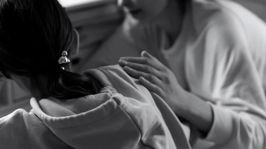 Black-and-white close-up of a person in a hoodie receiving a gentle hand on the shoulder, conveying quiet comfort, care, and emotional support.