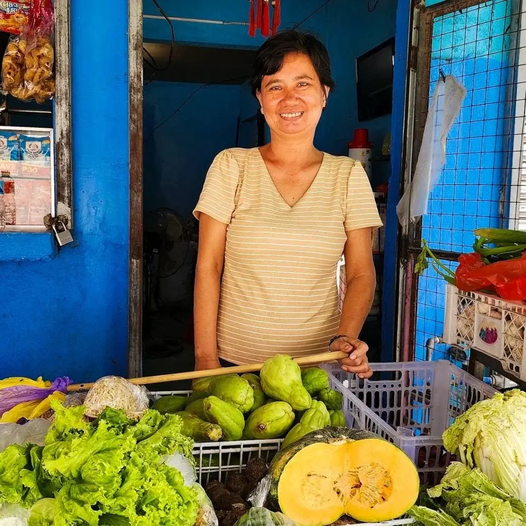Smiling market vendor stands behind a colorful stall of lettuce, melons, cabbage, peppers, and other fresh produce in a blue shop.