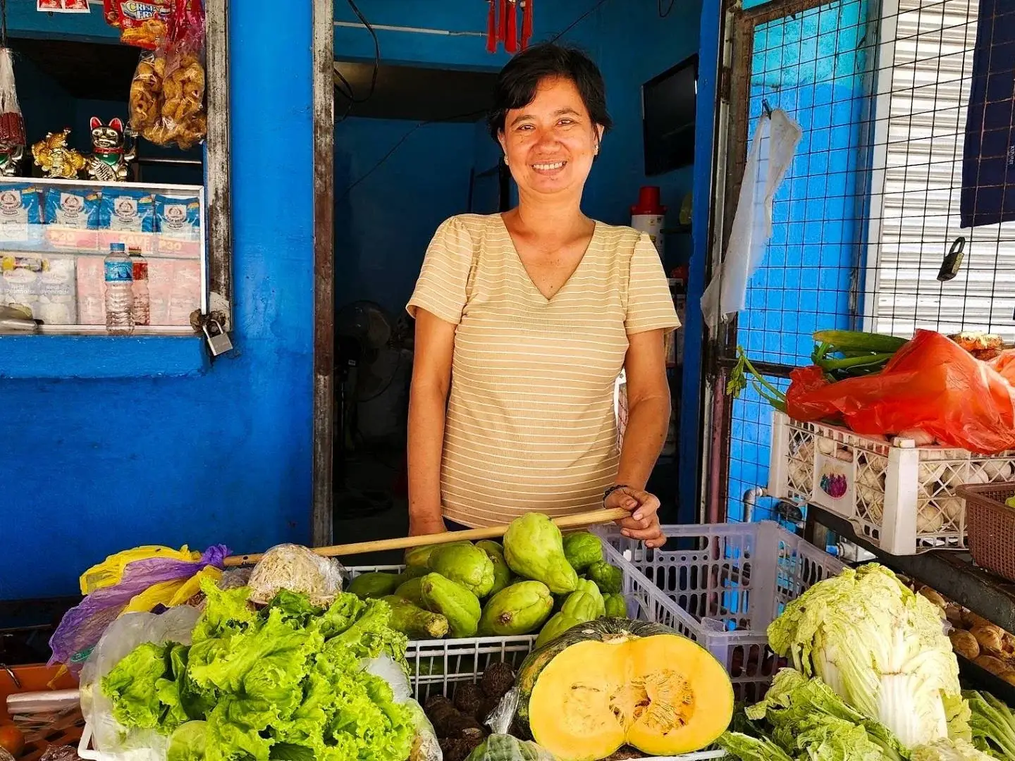 Smiling market vendor stands behind a colorful stall of lettuce, melons, cabbage, peppers, and other fresh produce in a blue shop.