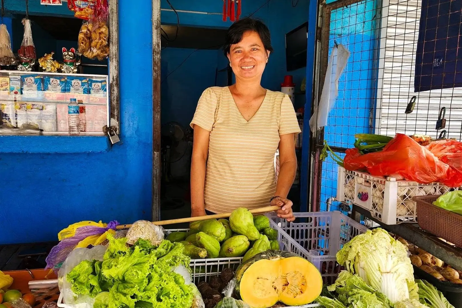 Smiling market vendor stands behind a colorful stall of lettuce, melons, cabbage, peppers, and other fresh produce in a blue shop.