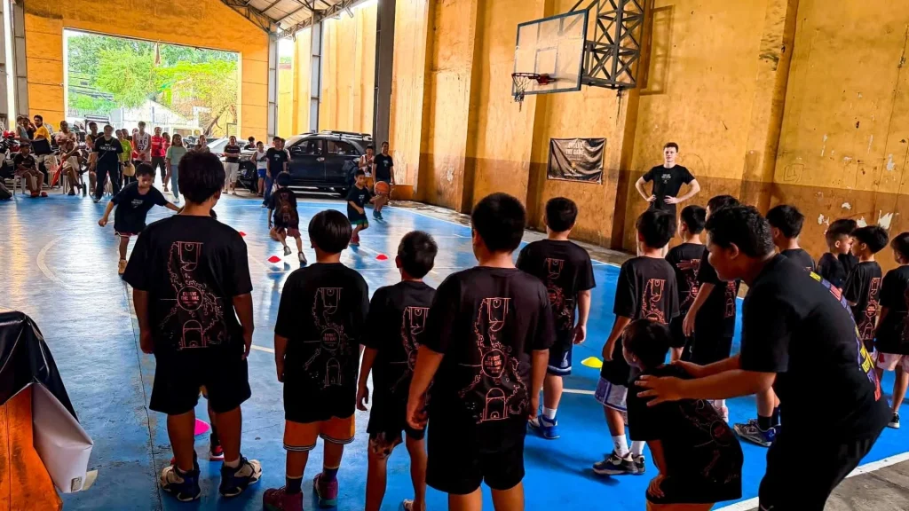 Filipino children in black sports uniforms gather on a blue indoor court as a coach addresses them near a basketball hoop; spectators watch in a bright gym.