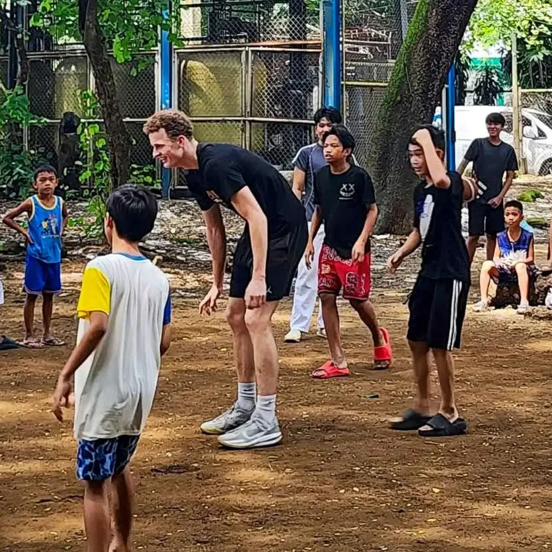 Adults and children gather in a lively urban park, playing outdoors in the Philippines as a tall man talks with a boy; trees, fences, and onlookers surround them.