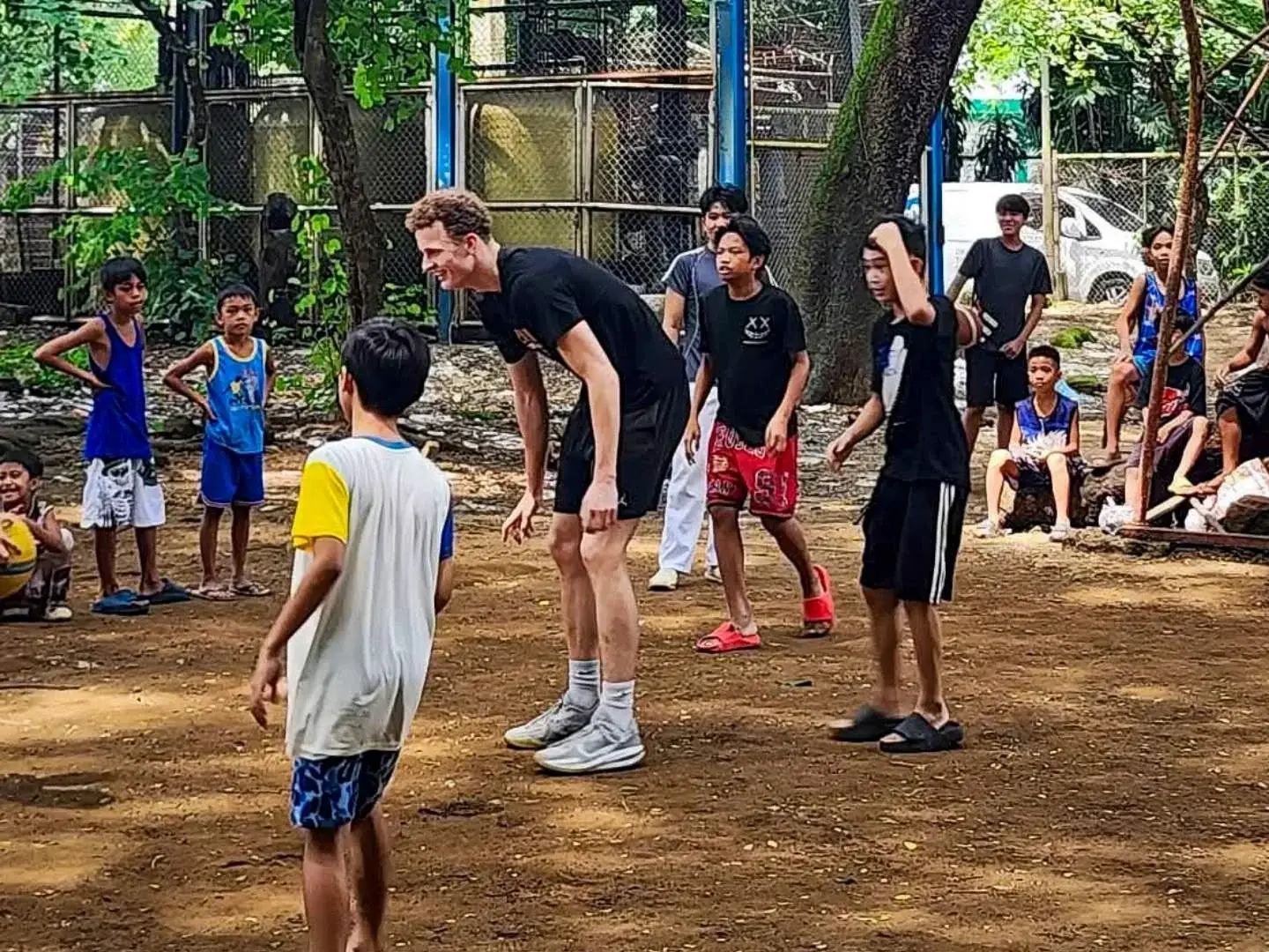 Adults and children gather in a lively urban park, playing outdoors in the Philippines as a tall man talks with a boy; trees, fences, and onlookers surround them.