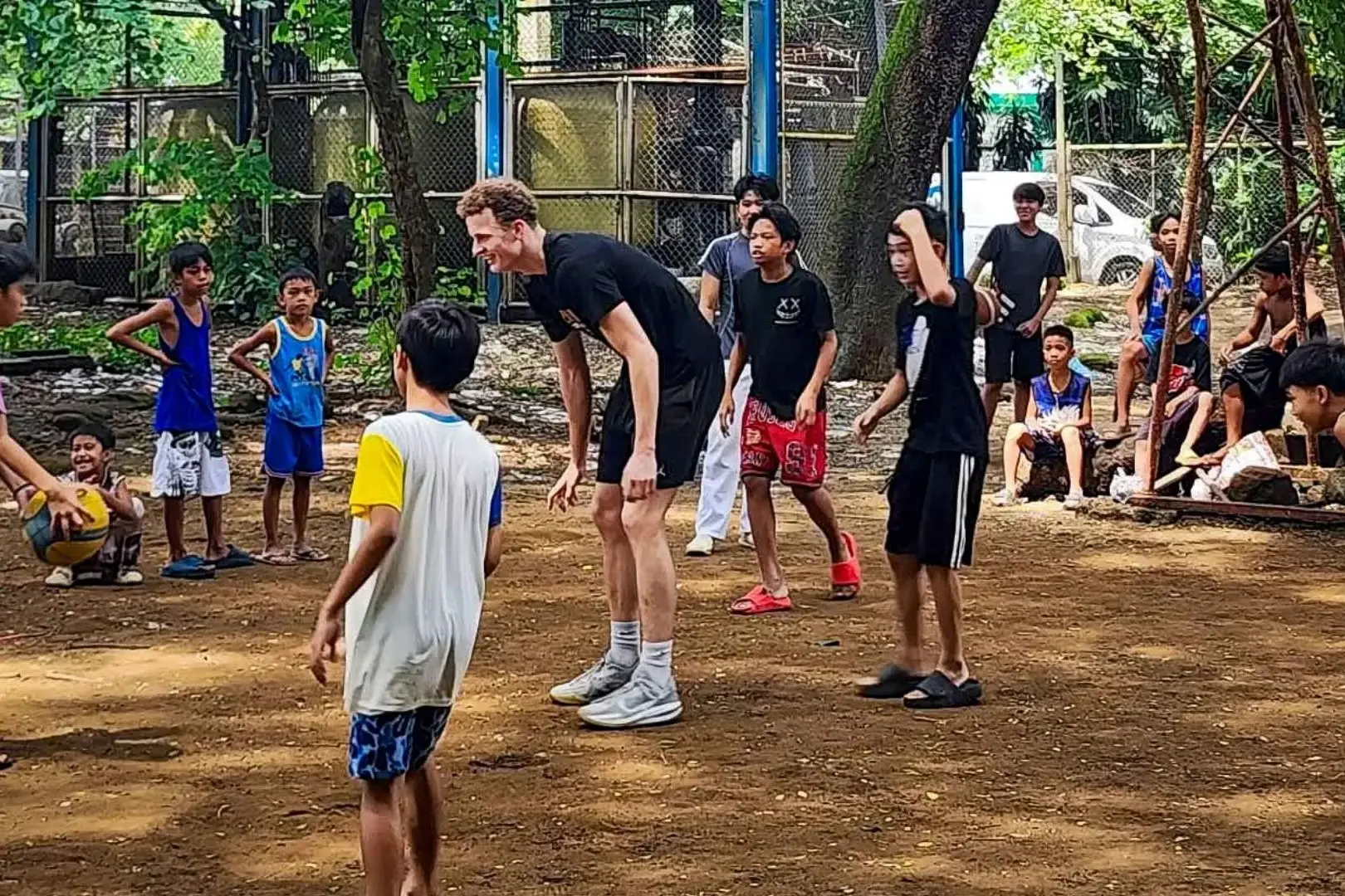 Adults and children gather in a lively urban park, playing outdoors in the Philippines as a tall man talks with a boy; trees, fences, and onlookers surround them.