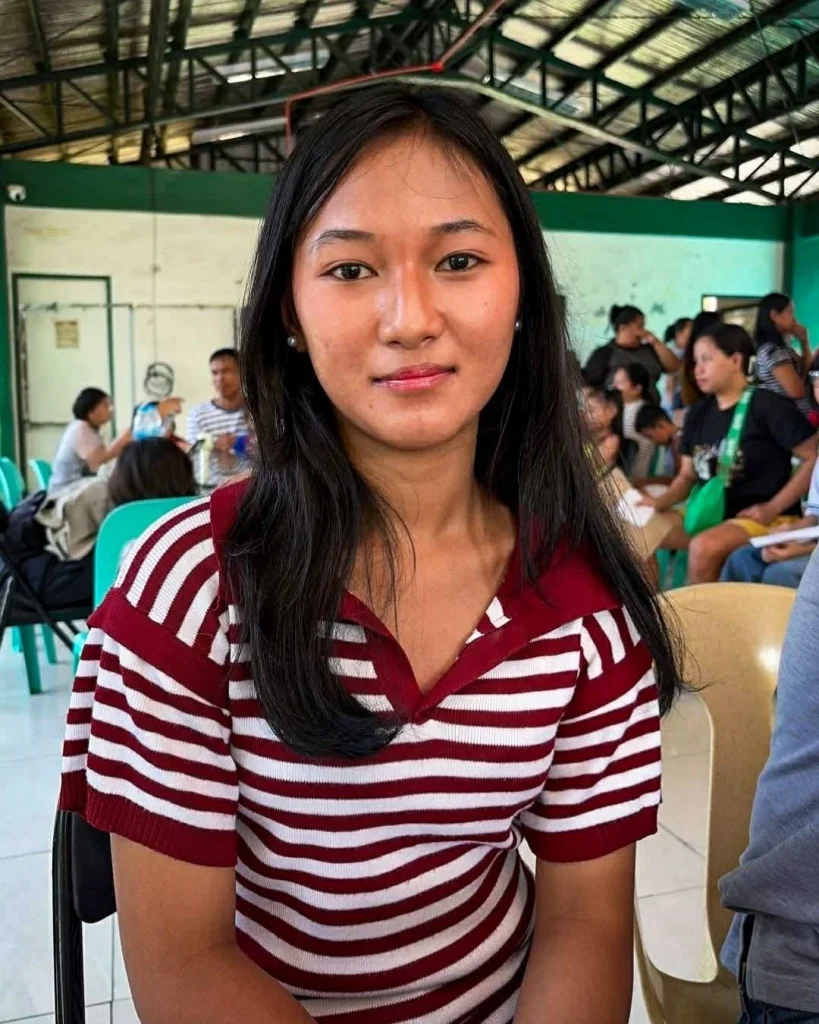 A young Filipino woman with long black hair in a red and white striped shirt sits indoors, with several people seated and talking in the background.