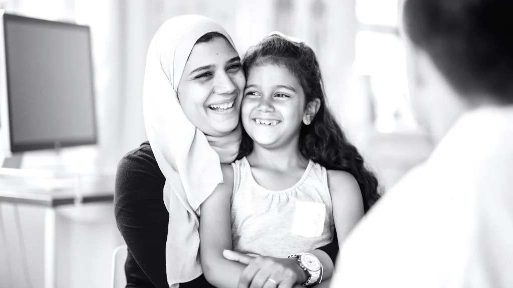Black-and-white photo of a smiling hijab-wearing mother hugging her young daughter, with a monitor and blurred person in the background.