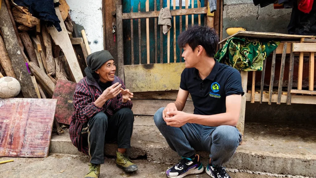 An elderly woman in a headscarf and striped jacket talks to a young man in a black polo shirt while they sit together on a concrete outdoor step.