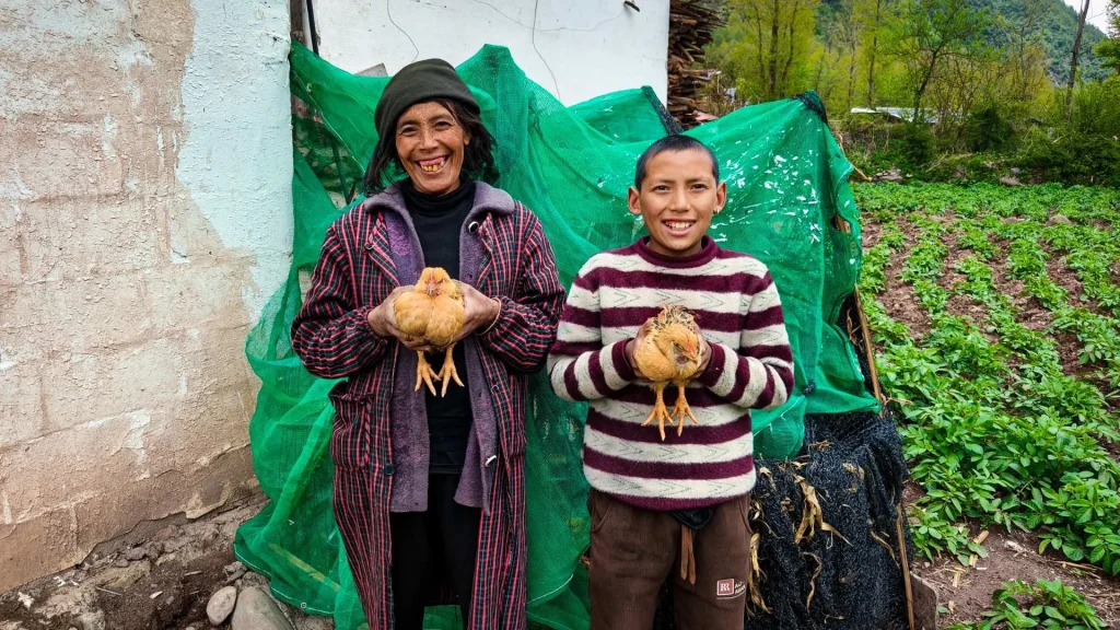 A smiling woman and boy hold chickens beside a rural garden, with green netting and crops behind them; the scene feels warm and welcoming.