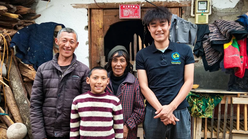 Four family members smile outside a weathered rural home, surrounded by stacked firewood, hanging clothes, and a wooden porch, creating a warm, welcoming scene.