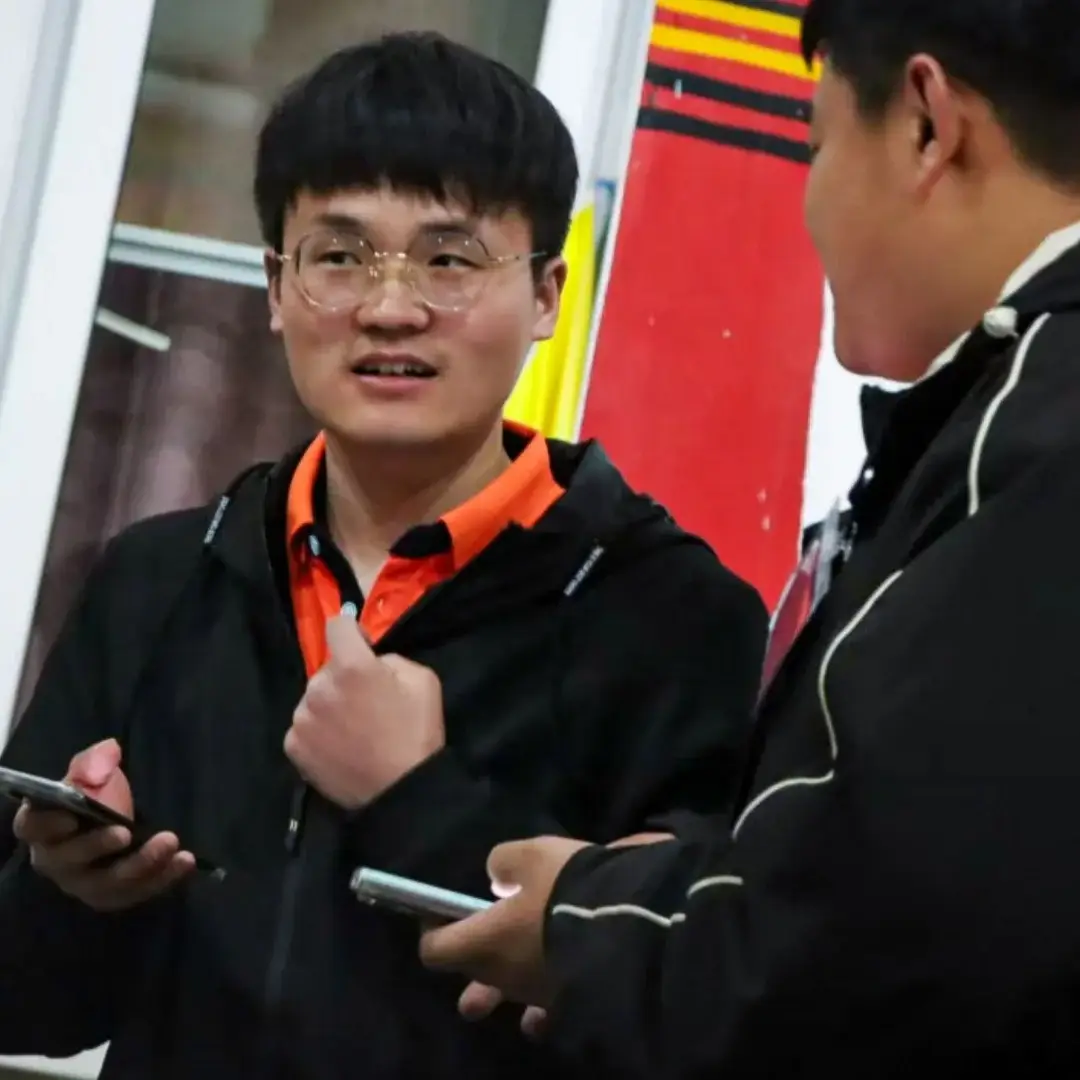 Two young men in black jackets are having a conversation; one wears glasses, holds a smartphone, and gestures while the other listens.