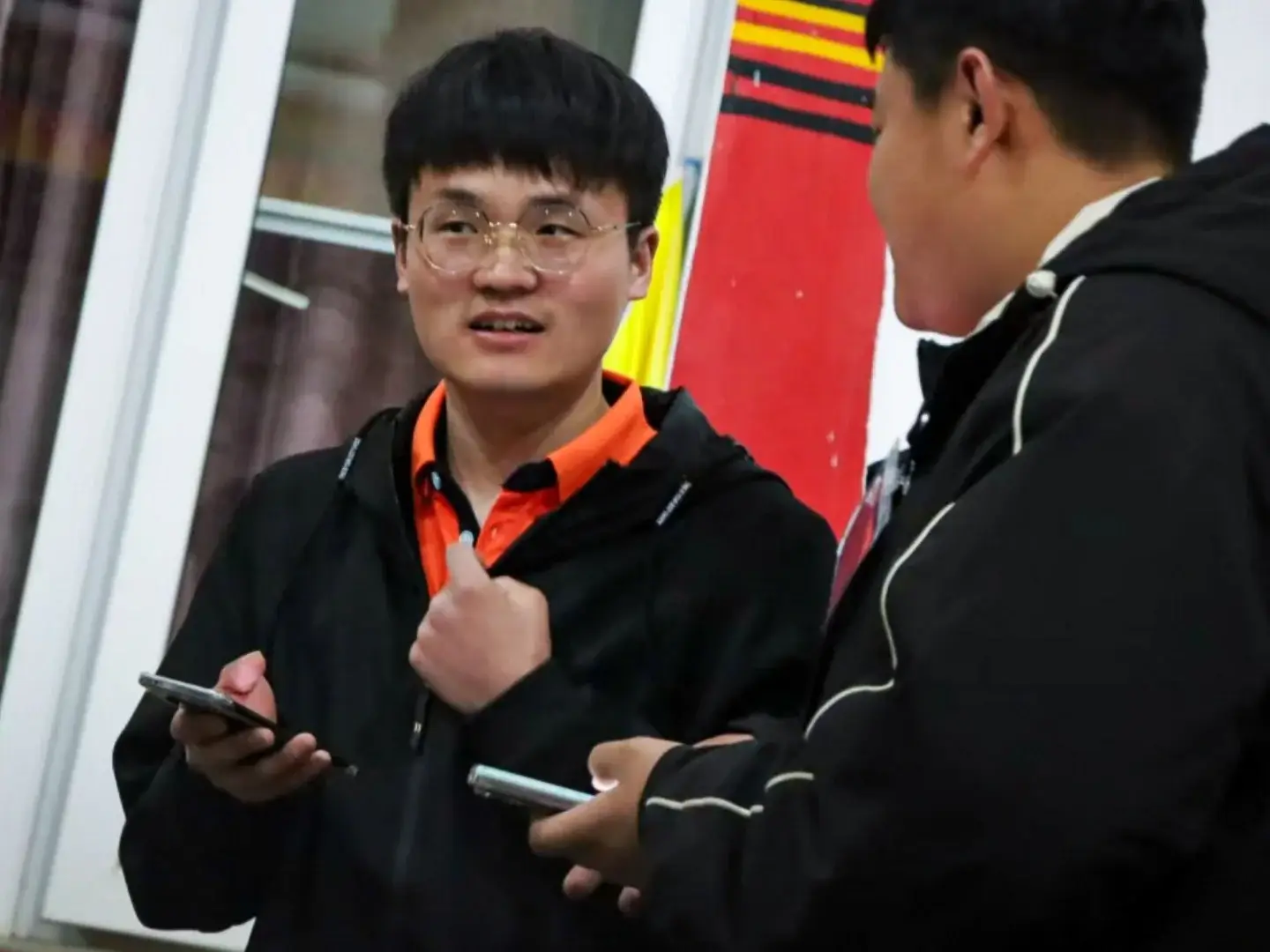 Two young men in black jackets are having a conversation; one wears glasses, holds a smartphone, and gestures while the other listens.