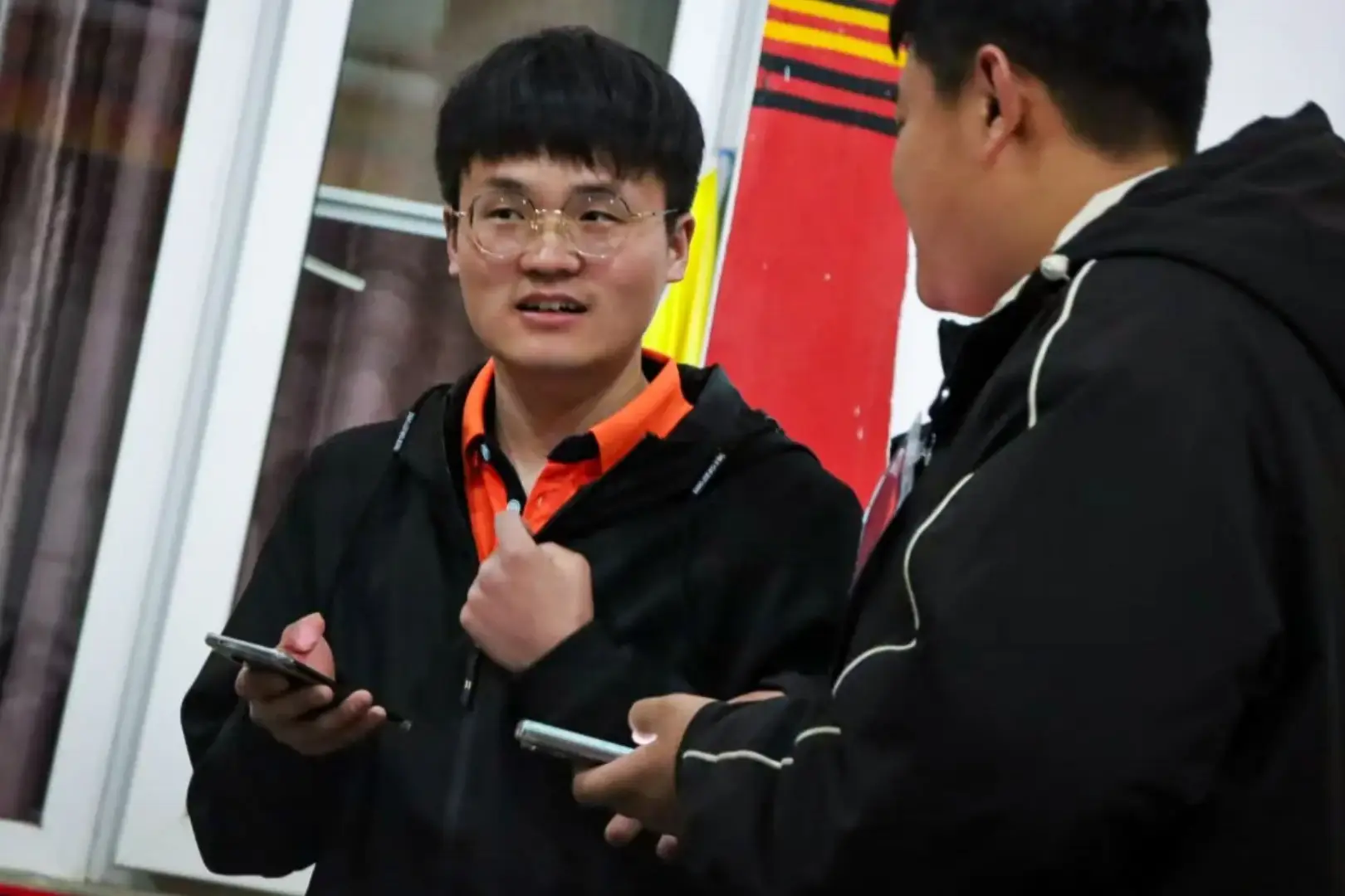Two young men in black jackets are having a conversation; one wears glasses, holds a smartphone, and gestures while the other listens.